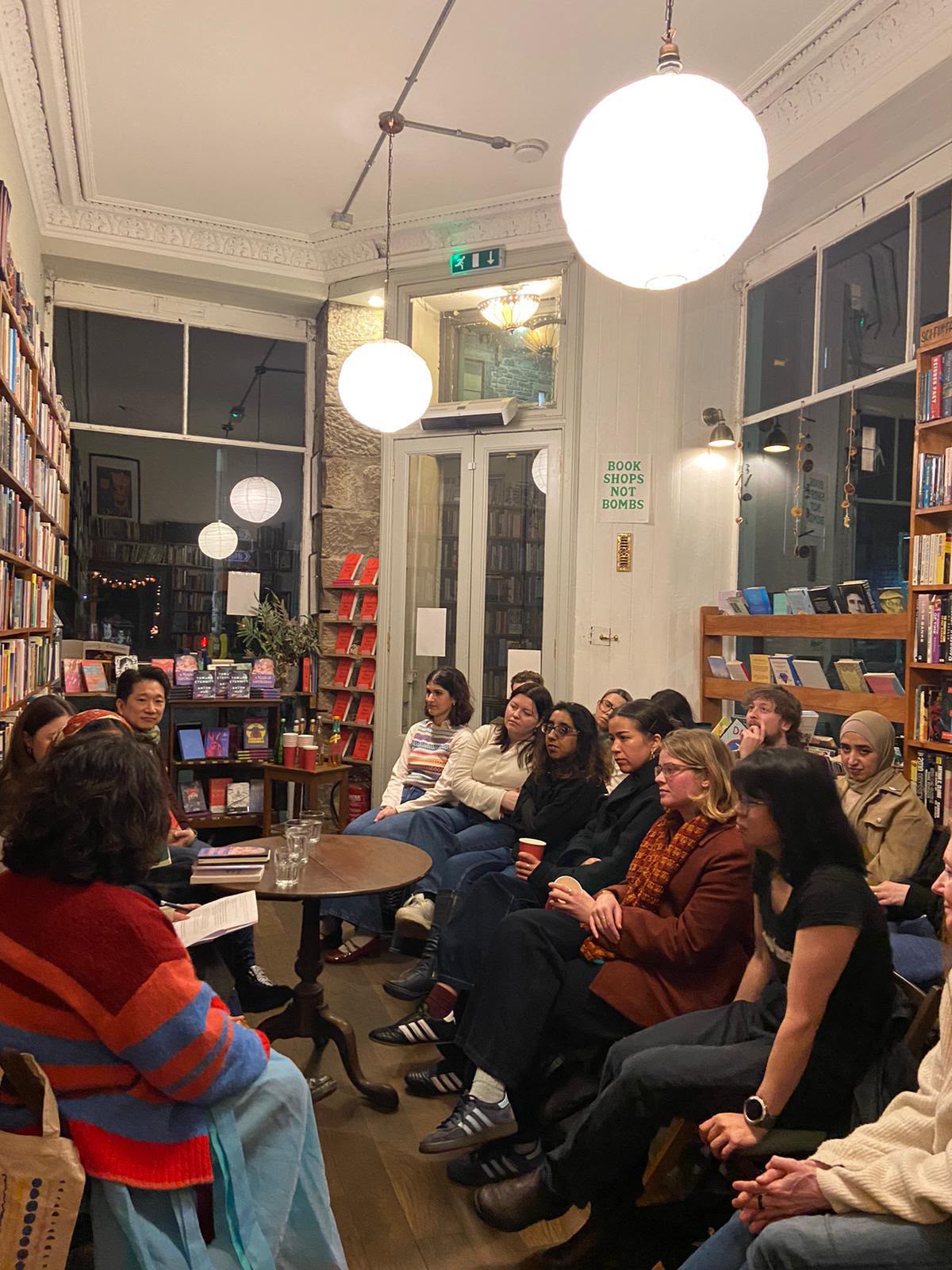 A full audience seated among the shelves at Tills Bookshop on a winter evening, lit by globe lamps, with a ‘Book Shops Not Bombs’ sign on the wall behind